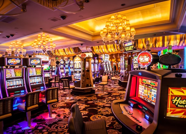 A vibrant casino interior filled with rows of colorful slot machines, each illuminated with bright lights and displays. The ceiling is adorned with elegant chandeliers and golden drapes, creating a luxurious atmosphere. The patterned carpet adds to the elaborate design of the space.